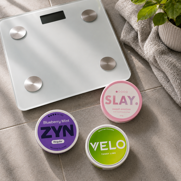 Bathroom scales on a tiled floor alongside a plant and towel with Slay, ZYN and VELO nicotine pouch cans resting in front of the scales.